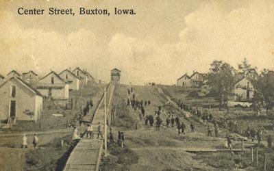 Many people seen at a distance walking down “Center Street” in Buxton, Iowa, circa 1908.  Photograph shows company houses, muddy streets, “coal chute hill,” and many mature trees.