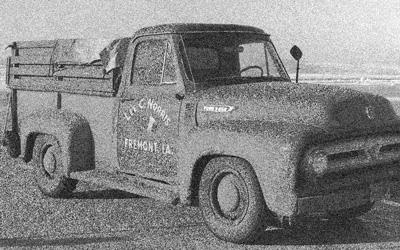 1950s era Ford F-250 truck with “Lee C. Norris, Freemont, IA.” painted on passenger’s door, is parked at the Des Moines airport awaiting the flight of 39 hogs from Iowa  to Yamanashi, Japan.  Back of truck is boarded up on the sides and covered with a strapped-down tarp.