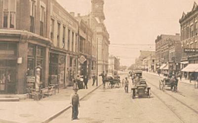This image shows a panoramic view of an intersection in downtown Fort Dodge. The streets are lined with 2-3 story brick buildings. A few people are walking on the sidewalks. Several horse-drawn buggies are in the street. A couple of cars are driving down one of the streets, and the tracks for a tram run down the middle of one street.