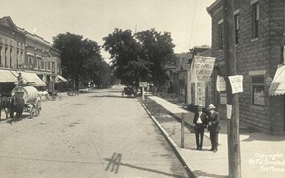 This image shows a panoramic view of an intersection in the business district of Iowa City. A few pedestrians are standing on the sidewalks, and several horse-drawn buggies are tied at hitching posts. The street is lined with 2-3 story brick buildings. Electrical poles are present on both sides of the center street and on one side of the street to the left. Each pole carries several electrical wires.