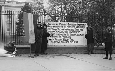 In this 1918 photograph taken by Harris & Ewing, two suffragettes hold a banner outside the gates of the White House accusing President Woodrow Wilson of hypocrisy when he claims to fight for and support democracy during World War I, all the while denying women the right to vote in his own country.