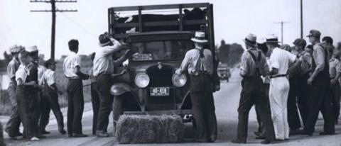 The photo depicts a bale of hay in the middle of the road blocking a truck with cows in the back bound for Sioux City with protestors on either side of the truck. 