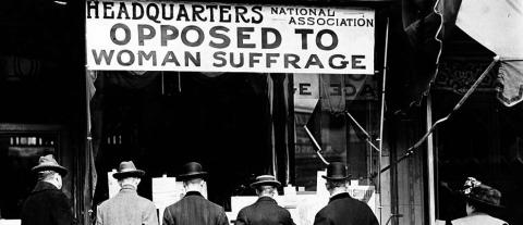 Photograph shows men looking at material posted in the window of the National Anti-Suffrage Association headquarters while a woman looks on behind them. The sign in the window reads "Headquarters National Association Opposed to Woman Suffrage."