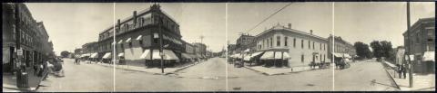 This image shows a panoramic view of an intersection in the business district of Iowa City. A few pedestrians are standing on the sidewalks, and several horse-drawn buggies are tied at hitching posts. The street is lined with 2-3 story brick buildings. Electrical poles are present on both sides of the center street and on one side of the street to the left. Each pole carries several electrical wires.