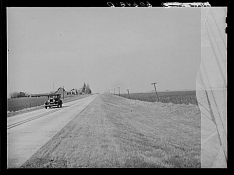 Image of a car driving through Grundy County, Iowa.  The image shows a farm in the background and the road is passing through fertile farmland.