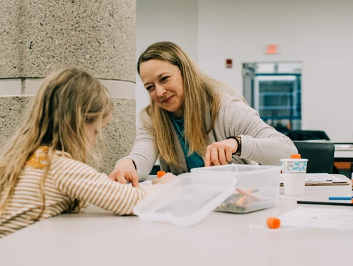 A woman smiles and helps a little girl with a workshop activity