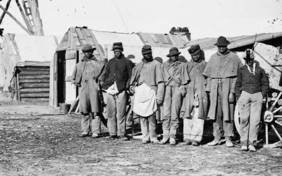 One of the Civil War photographs compiled by Hirst D. Milhollen and Donald H. Mugridge, this 1864 image depicts seven “contraband” teamsters dressed in old Union uniforms standing near a wagon and shack.  