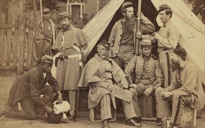 Between 1861 and 1865 George N. Barnard and C. O. Bostwick photographed this group of white soldiers, possibly the 8th Company, 7th New York Infantry, at Washington D.C.’s Camp Cameron. An African-American “contraband” kneels to the left of the group.