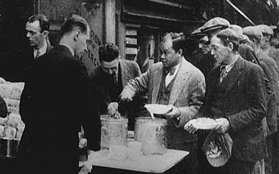 The black and white photo shows a long line of men waiting in a bread line.  