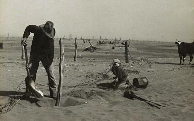 The image shows a farmer digging up a fence and attempting to move it higher to prevent it from being buried in the dust that is accumulating around it.  There is a young boy on his knees close by.