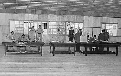 Three tables are seen in a plain looking room with military personnel seen through the background windows.  General Harrison is seated at the table on the left, General Nam is seated at the table on the right.  Both Generals are attended by other staff as they sign the armistice that ended the three year Korean conflict. 