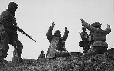 In a grassy hill area, two men are seen kneeling with their arms in the air. Three soldiers are seen on each side of them with guns.  All men are dressed in cold weather gear.