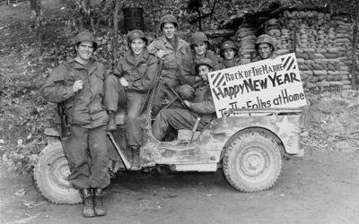 Seven uniformed, smiling, relaxed soldiers from Iowa are seen seated on an army jeep. They hold a sign that reads: “Rock of the Marne • Happy New Year • To The Folks at Home.” The background is a wooded hillside with a large amount of sandbags stacked up.