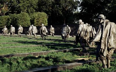 Multiple statues visible in an are of low-lying bushes and granite blocks.  United States flag is seen on the left.  Mature trees are seen in the background.