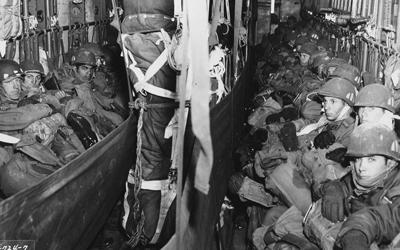 Photo of inside of a cargo plane soldiers lining the two sides of the cargo hold.  Parachute equipment seen in the center hanging from ceiling of the cargo hold.