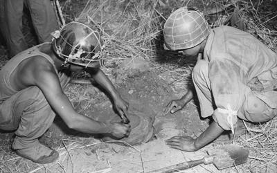 Two men seen squatting down, leaning over circular object they are in the process of burying in the ground.  Each man wears a helmet but no other uniform or safety equipment.  A third man is seen standing behind the man on the left along with a pile of straw in the background.