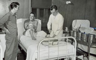 Three men seen in Great Lakes Hospital in Korea.  One man sits smiling on a hospital bed while another man bandages his left foot.  A third man is seen talking with the injured man.  Medical equipment sits on a cart near the man who is doing the bandaging.