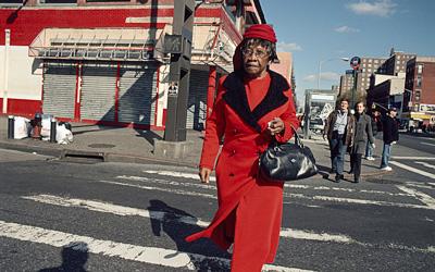 Photograph shows a woman in a bright red coat with matching hat, walking in a crosswalk.