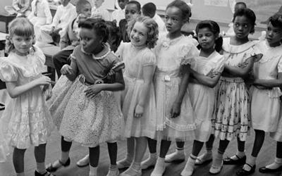 Photograph shows a line of African American and white school girls standing in a classroom while boys sit behind them.
