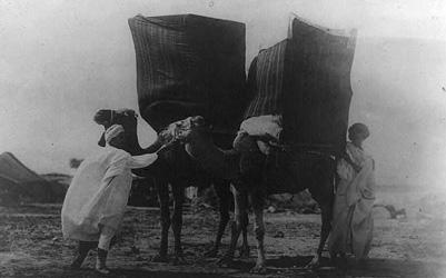 Image shows two men in the Sahara with very large wares bags on the backs of two camels. 
