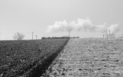 Image of Iowa land that has been freshly plowed on one side of the image, and not plowed on the other.  There is a train and telephone poles in the background.