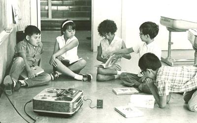 This photograph shows children – Alberto Paz, Virginia Guzman, Dominga Castillo, Mario Castillo, and Junior Castillo – listening to recordings of their voices on a tape recorder. Each of the kids reads portions of a story to have their voices recorded. They were enrolled in the intermediate school program for migrant children. The goal of the Migrant Education Program was to ensure that all migrant students reach challenging academic standards and graduate with a high school diploma (or complete a GED).
