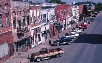 Landscape photograph of main street with multiple buildings visible along with sidewalks, lampposts, trucks, and cars.
