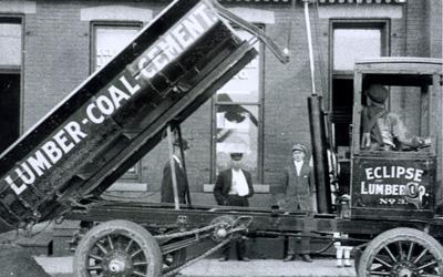 Old-fashioned delivery truck with box hoisted in the air.  “Lumber - Coal - Cement” printed on the side of the box, and four men are also seen in the photo.