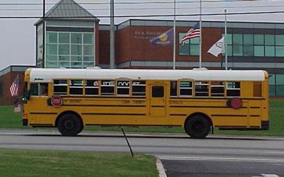 Large, modern school bus driving on street pictured with school building in background.