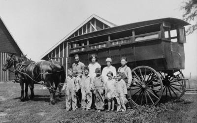 Large, horse-drawn wagon with four older and six younger students posed in front of it.  “Webster Consolidated School District” is painted on the side of the wagon.