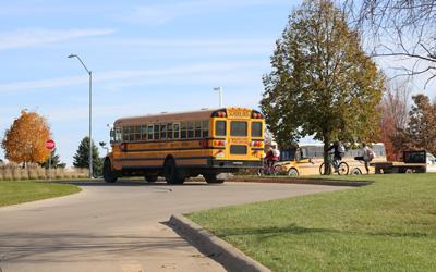 Two school busses in Pella, Iowa, 2018.  One bus is about to pull out onto the street and the other bus is about to turn into the school drive.  Two kids riding bicycles and another student standing on the sidewalk are also visible.