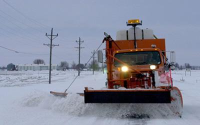 Large, orange snowplow removing snow on a paved road somewhere in rural Iowa.  The only person in the photo is the driver of the snowplow.