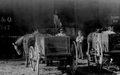 Two horse-drawn wagons parked near train cars.  Doors are open on train cars, and three men and a child are also seen in the photo.