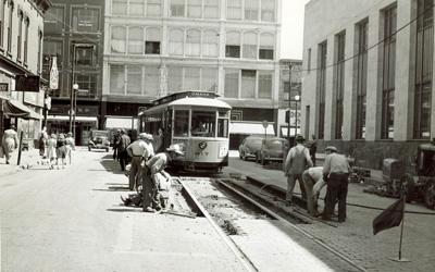 Parked streetcar seems to wait for workers to finish repairs to rails and roadway in front of it.  Four story buildings seen in the background, and two story buildings on either side of the streetcar along with parked cars and a few pedestrians.