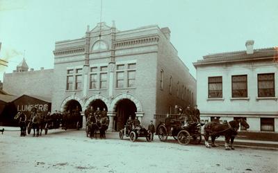 Fire Station Number One is an ornate brick building with three horse-drawn fire wagons and one early automobile firetruck all displayed in the front of the building along with firefighters.