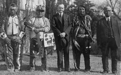 President Calvin Coolidge is seen in the center of a group of five Native American men on the south lawn of the White House.