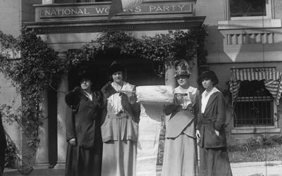 Four women in dresses and hats proudly stand in front of a building with a sign that reads “National Women’s Party.”  Between the two women in the center is a very large roll of paper that the women are holding up.  These National Women’s Party leaders had been working to collect signatures of support on a “mile long” paper to present to members of Congress.