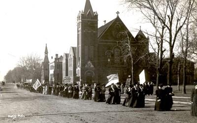 Women's suffrage parade shown passing by church. One banner reads: "Taxation without representation is tyranny--as true now as in 1776". 