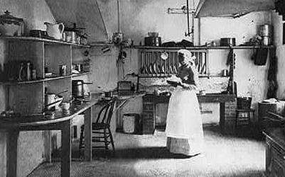 One adult woman in a floor-length dress, seen in a large kitchen.  Commercial range and oven on the right, many shelves, tables, and other cooking equipment also visible.