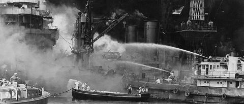 The black and white image shows the aftermath of the bombing of Pearl Harbor, Hawaii with the  USS West Virginia aflame. Disregarding the dangerous possibilities of explosions, United States sailors man their boats at the side of the burning battleship, USS West Virginia, to better fight the flames started by Japanese torpedoes and bombs. Note the national colors flying against the smoke-blackened sky