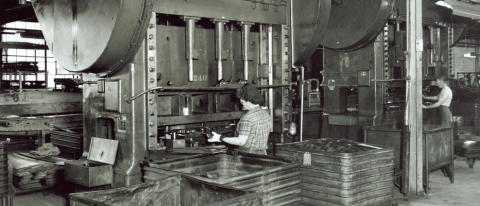 Woman in foreground standing with her back to the photographer, operating a very large boring press.