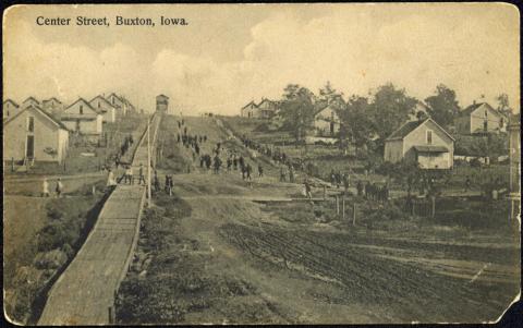 Many people seen at a distance walking down “Center Street” in Buxton, Iowa, circa 1908.  Photograph shows company houses, muddy streets, “coal chute hill,” and many mature trees.