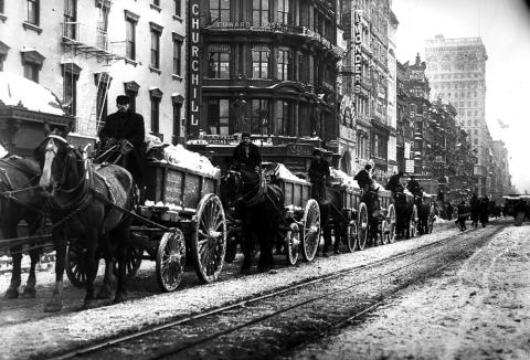 This image shows a line of several horse-drawn wagons hauling snow down a  New York City street. 