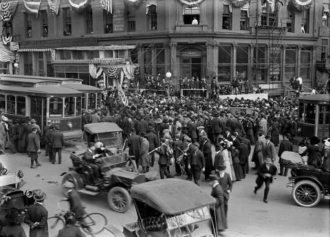 A crowd gathers on a busy intersection in Washington, D.C. and trolley cars, automobiles, a bicycle are shown in the photograph. 