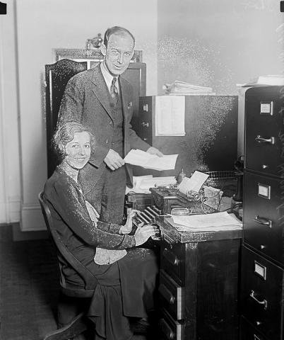 A man and a woman pose in an office with a typewriter.