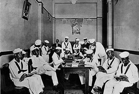 A photo of African American soldiers and sailors sitting and standing around a table in a Red Cross Rest Room specifically outfitted for only African Americans during World War One.  This photo was taken at the Red Cross headquarters, Branch Number 6, of the New Orleans Chapter.