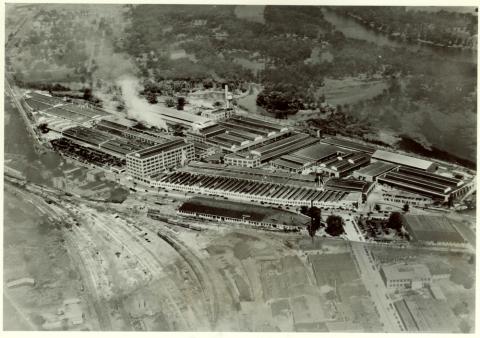 Aerial photograph of a large manufacturing facility.  More than fifteen buildings are visible within the complex, and it even has two water towers.  A network of railroad lines are in the foreground, and a river is in the background.