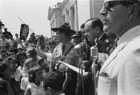 Photograph shows a group of people, one holding a Confederate flag, surrounding speakers and National Guard, protesting the admission of the "Little Rock Nine" to Central High School.