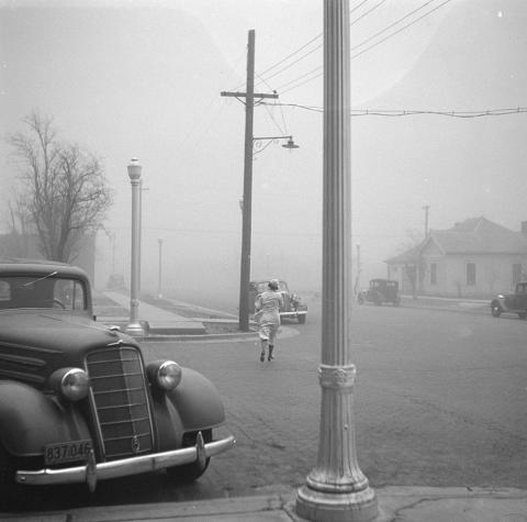 Image showing a parked car in the foreground of a town with a woman wearing a dress holding her hat on her head.  A dust storm is engulfing the town.