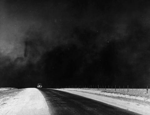 The image shows a very heavy black cloud of dust behind a single car that is driving on a country road.